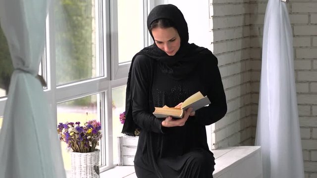 Beautiful meddle eastern young woman reading book next to window.