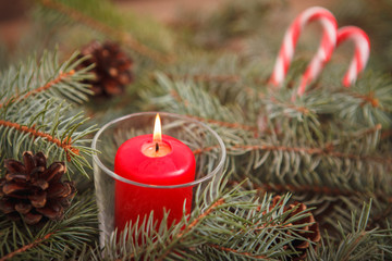 New Year composition, burning red candle in a candlestick on spruce branches with cones on a wooden background with candy-cane. Close-up. Christmas.