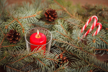 New Year composition, burning red candle in a candlestick on spruce branches with cones on a wooden background with candy-cane. Close-up. Christmas.