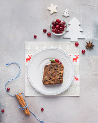 A piece of traditional Christmas English pudding (fruit cake) on white plate on a concrete light background. Christmas decor of the table. Funny napkins with reindeers Santa Claus.  Flat lay, top view
