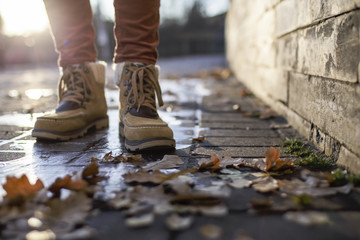 Legs in boots stand on a cobblestone pavement made of stone in a puddle covered with ice, in the rays of the sun, around are fallen leaves. Close-up.