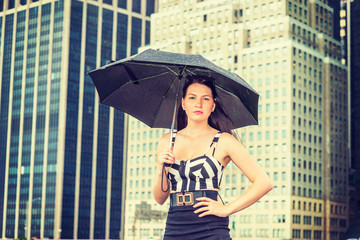 Fototapeta premium Raining day - grainy, foggy, drizzly feel. American Woman wearing patterned crop bra top, black skirt, sunglasses on head, holding umbrella, standing in business district in New York, looking at you..
