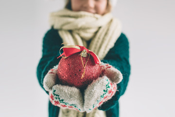 Big red Christmas ball in hands at the girl. The child is dressed in sweater, christmas hat and scarf studio shot .