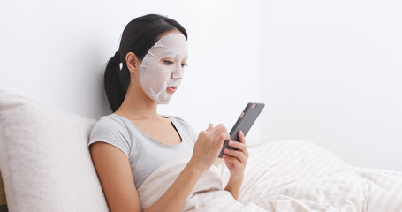 Woman apply facial mask on bed and reading on cellphone