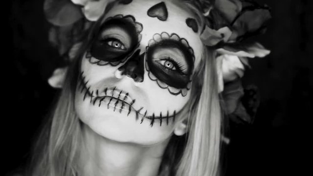 Closeup face of woman with Mexican sugar skull makeup and flowery wreath looking into the camera. Creative, artistic, Halloween concept - black and white slow motion video