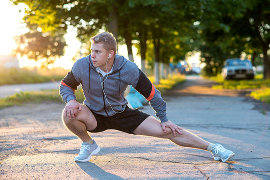 A Young Guy Runner In The Early Morning Makes Gymnastics Of His Feet In Headphones. A Warm-up Of The Joints Of The Muscles, A Man In The Park Listening To Music, A Confident Look. In Summer, Life