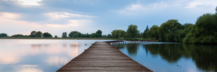 Wooden pier, bridge over lake at dramatic sunset with stormy clouds. Kuchyna, Slovakia. Panorama 3:1