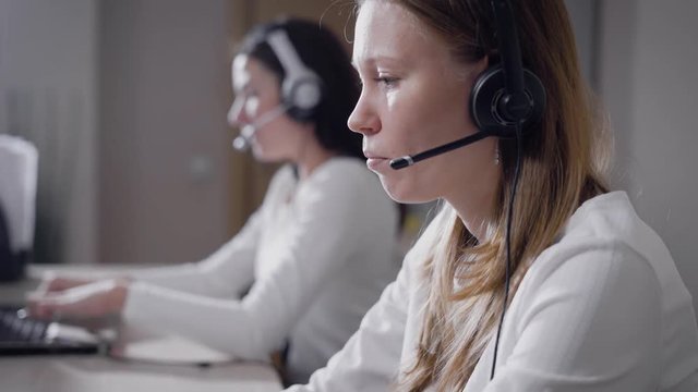 employees women are sitting at tables with headphones with a microphone on heads, doing outbound calls and typing on a keyboards of a laptops