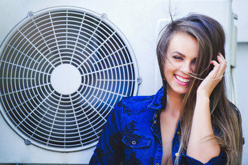 Beauty young woman with air conditioning. near the fan happy laughing. Long hair. Fashionable lady with a beautiful hairdo, makeup. against the background of the outdoor air conditioner