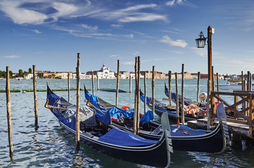 Fototapeta na wymiar Blue gondolas in Venice moored to the jetty between wooden piles with view of the sea and the palace at sunset
