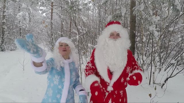 Santa in a red hat and with a white beard and a beautiful merry granddaughter make an air kiss on the camera in a snowy forest. Slow Motion Picture