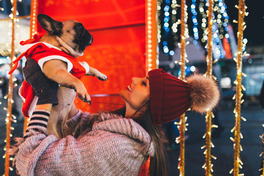 Beautiful young brunette woman with French bulldog enjoying Christmas or New Year night on a city street. 