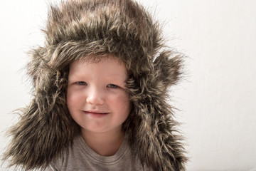 Happy child playing at home. The funny guy put on a warm fur hat and admires himself. Close-up. Portrait with emotions.