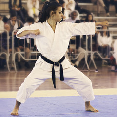 A black belt girl is performing a kata at a karate competition. People out of focus in background.