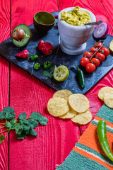 Mexican food concept:homemade guacamole, tortilla chips, salsa, chilli and fresh ingredients over vintage red rustic wooden background. 