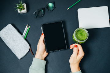 Tablet in woman hands and Jar with fresh green smoothies at office desk on black table.