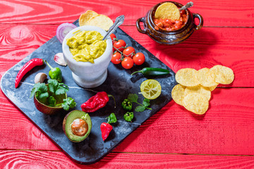 Mexican food concept: homemade guacamole, tortilla chips, salsa, chilli and fresh ingredients over vintage red rustic wooden background. 
