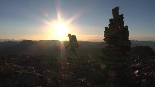 Man walks past rock stack on mountain top as sun begins to set.