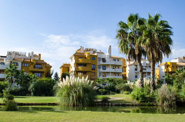A small park with palm trees, bushes, tall grass and a shaved green lawn in a residential area with multi-storey yellow Spanish houses of the Punta prima in Torrevieja, Spain. A great place for summer