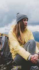 Hipster young girl with backpack hiking in mountains. Tourist traveler sitting on the rocks and enjoying the view from the top of the mountain