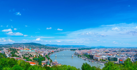 Obraz premium Panoramic cityscape view of hungarian capital city of Budapest from the Gellert Hill. The bridges connecting Buda and Pest across the river Danube. Summertime sunshine day, blue sky and green of trees