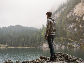 Handsome yung man standing on background of woods on shore of lake looking away.