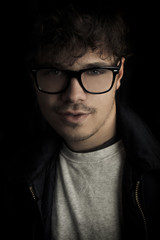 Fashion dark  portrait of sexy young man with glasses on black background. Portrait of intelligent man. Photo portrait of curly fashion model with glasses with dark black background. Vintrage shot.
