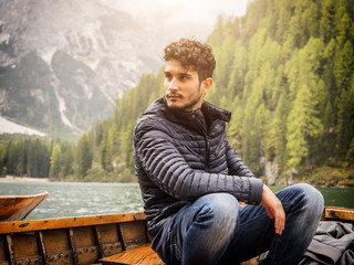 Fototapeta premium Handsome tattooed man sitting in boat on background of woods on shore of lake looking away.
