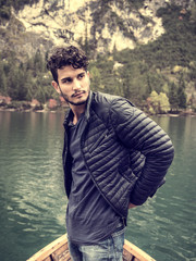 Fototapeta premium Handsome tattooed man sitting in boat on background of woods on shore of lake looking away.