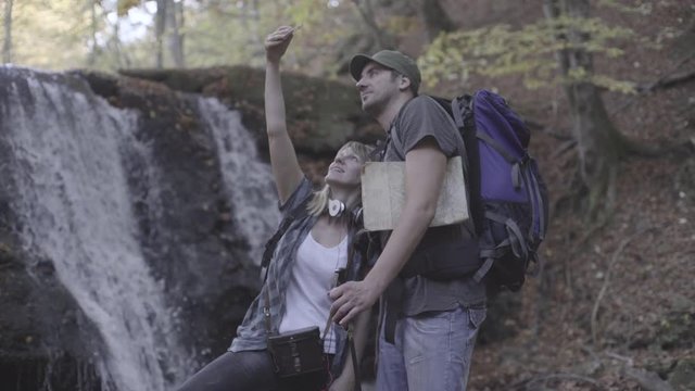 Happy young couple take photo, blond woman and man with cap smiling in camera while making portrait selfie with mobile phone during hiking, waterfall and forest in the blurred background, steady cam