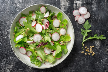 Healthy salad with radish, lettuce, arugula and pine nuts on black background. Top view. 