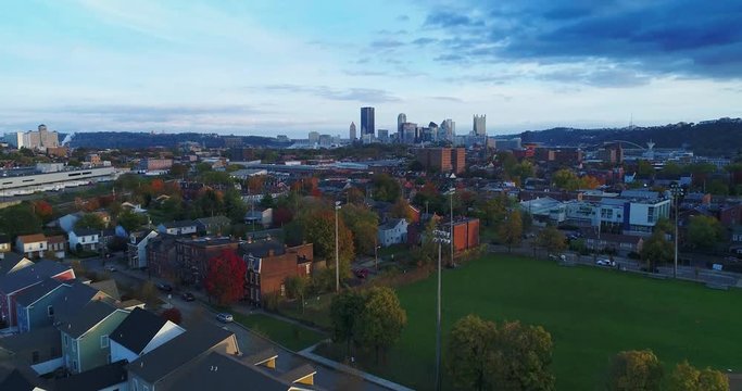 A late evening rising aerial establishing shot from a residential neighborhood with the Pittsburgh skyline in the distance. Shot at 48fps.  	