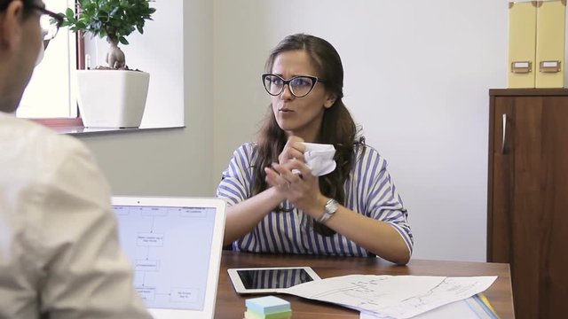 Female employee crumples papers, talking to colleague angrily in office. Young woman waving working documents then crush down them in clod while speaking to man sitting at table with laptop
