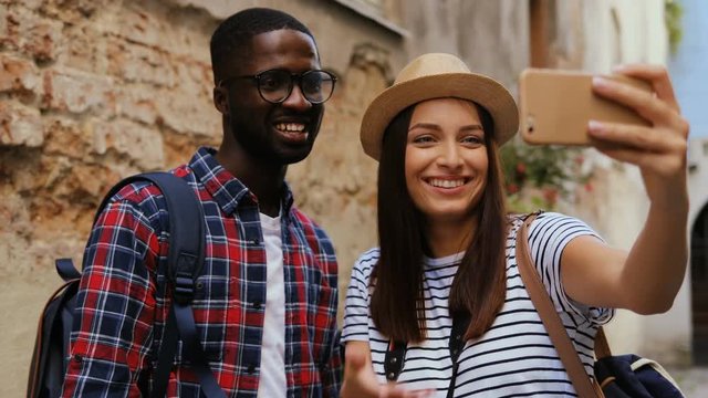 Beautiful cheerful young tourist couple of students video chatting on smartphone on vacation in old street of beautiful city. Sightseeing.