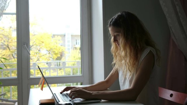 Young Caucasian girl in white t-shirt working in his house with his laptop near the window. Engaged in freelance print texts