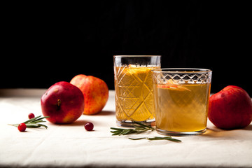 Two vintage glasses with apple cider on black background. Christmas beverages concept. Two red apples and rosemary sprig aside.  Warm backlight. Horizontal composition. Side view.