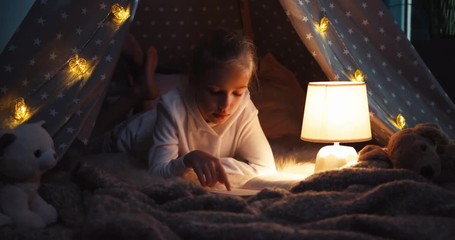 Portrait cheerful girl 9 years old reading book at night in the wigwam. Dolly shot