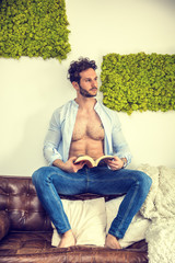 Shirtless sexy male model lying alone on couch in his living-room, looking at camera with a...