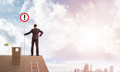 Businessman on house top showing sign with exclamation mark. Mixed media