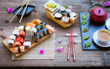 Fototapeta na wymiar Red teapot, green leaves, slate tray and red chopsticks on old wooden table, Sushi rolls, nigiri, raw salmon steak, rice, cream cheese, avocado, lime, pickled ginger, asian background, top view, 