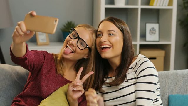 Two attractive female friends making funny selfies on the smart phone while sitting on the sofa in the modern living room. Indoor