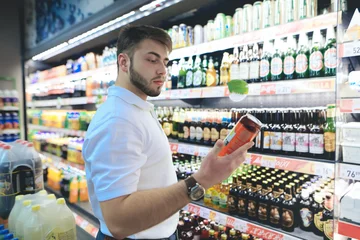 Zelfklevend Fotobehang A handsome bearded man chooses beer in a supermarket. The buyer buys alcohol at a supermarket. A man looks at a can of beer © bodnarphoto