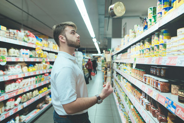 A beautiful man chooses canned food from supermarket shelves. A man with a beard juggles the goods in the store.