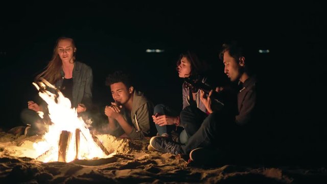 Multiracial group of young boys and girls sitting by the bonfire late at night and singing songs and playing guitar