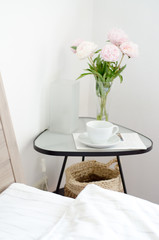 White bedroom in the morning; little glass table, lamp, basket,  white cup , bowl with pink peony and magazine