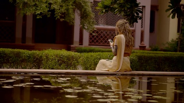 Girl devotee in kṛṣṇa consciousness is sitting in India on Board an artificial pond and writes SMS message on a mobile phone