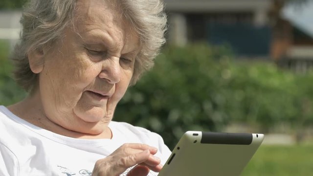 Mature old woman dressed t-shirt sitting on the background the green park outdoors in summer holds a silver computer tablet for looking the images. Slow motion