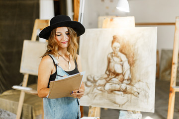 Beautiful long hair artist girl, in a black hat and denim dangarees, considering the idea of her picture before the easel