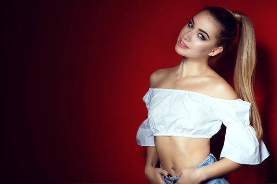 Closeup portrait of young beautiful blond lady with perfect make up and long ponytail wearing white short open shoulders top and blue jeans isolated on bright red background. Studio shot. Copy-space