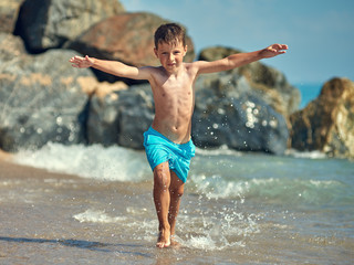 Cute Caucasian boy is running in the water along the sea shore against big boulders. His arms are wide open.